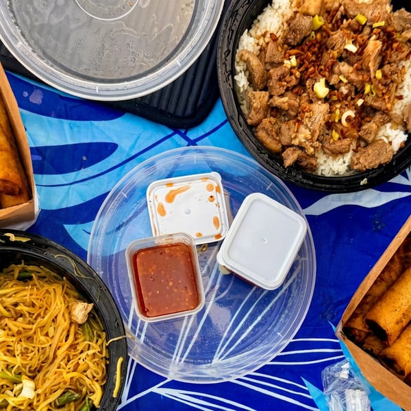 An overhead shot of a takeout meal with spring rolls, noodles, beef and rice, and sauces.